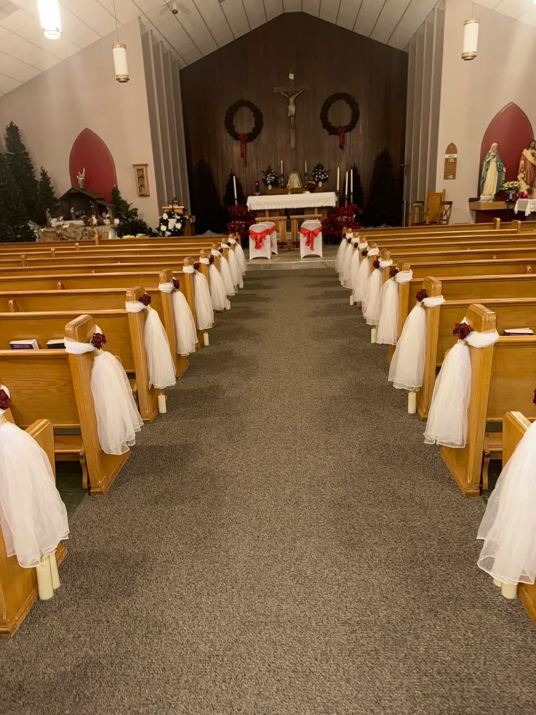 Church aisle with white tulle pew ends, burgundy roses, pillar candles, altar with Christmas wreaths and poinsettias