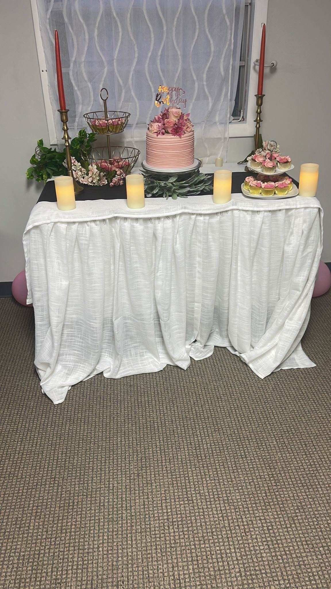 Pink birthday dessert table with floral cake, cupcakes, gold candlesticks, and soft candlelight